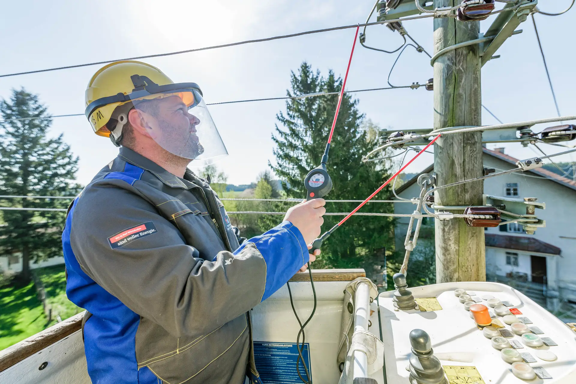 Ein Elektriker der Stadtwerke steht auf einem Hubsteiger und misst mit einem Spezialwerkzeug in Schutzausrüstung die Spannung an einem Holz-Strommasten.