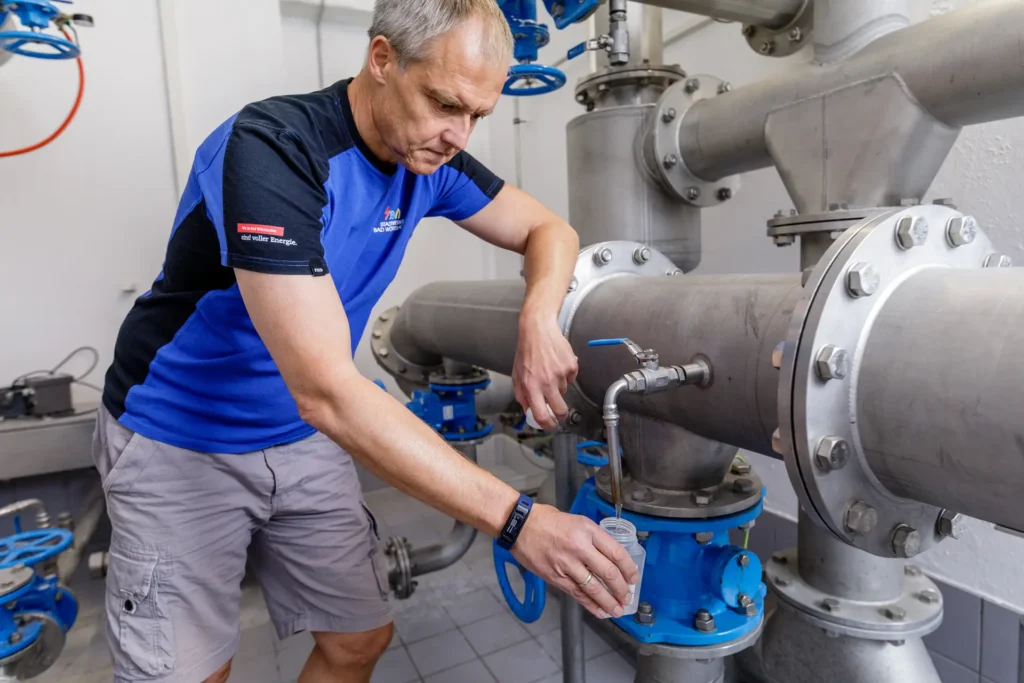 Ein Mitarbeiter der Stadtwerke entnimmt eine Wasserprobe im Technikraum eines Hochbehälters. Es sind große Metall-Wasserrohre zu sehen. 