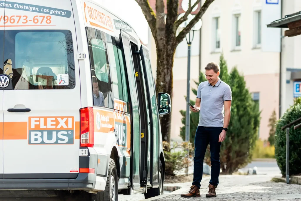 Junger Mann steht vor einem Flexibus und schaut auf sein Smartphone
