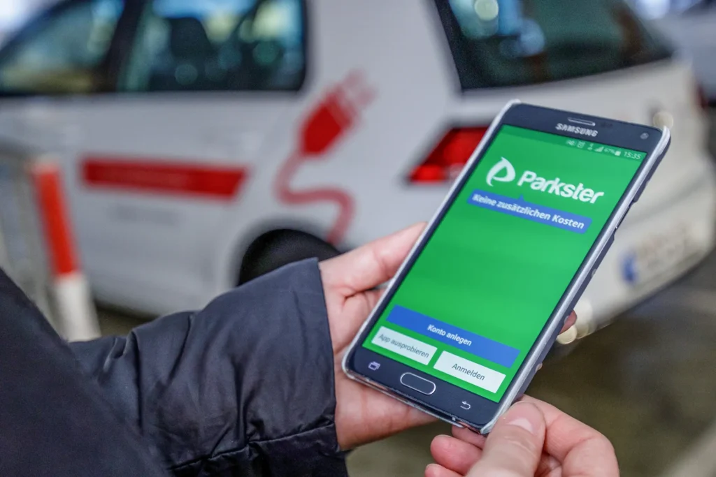 Eine Hand hält ein Smartphone in der Hand. Auf dem Smartphone ist die App "Parkster" installiert. Im Hintergrund ist unscharf ein Auto.