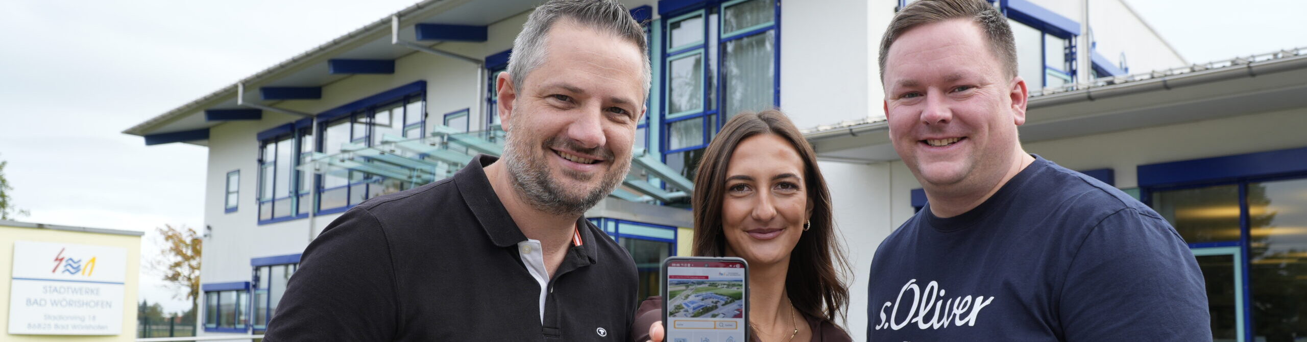 Drei Personen, die in die Kamera blicken. Die Person in der Mitte halt ein Smartphone in die Kamera.