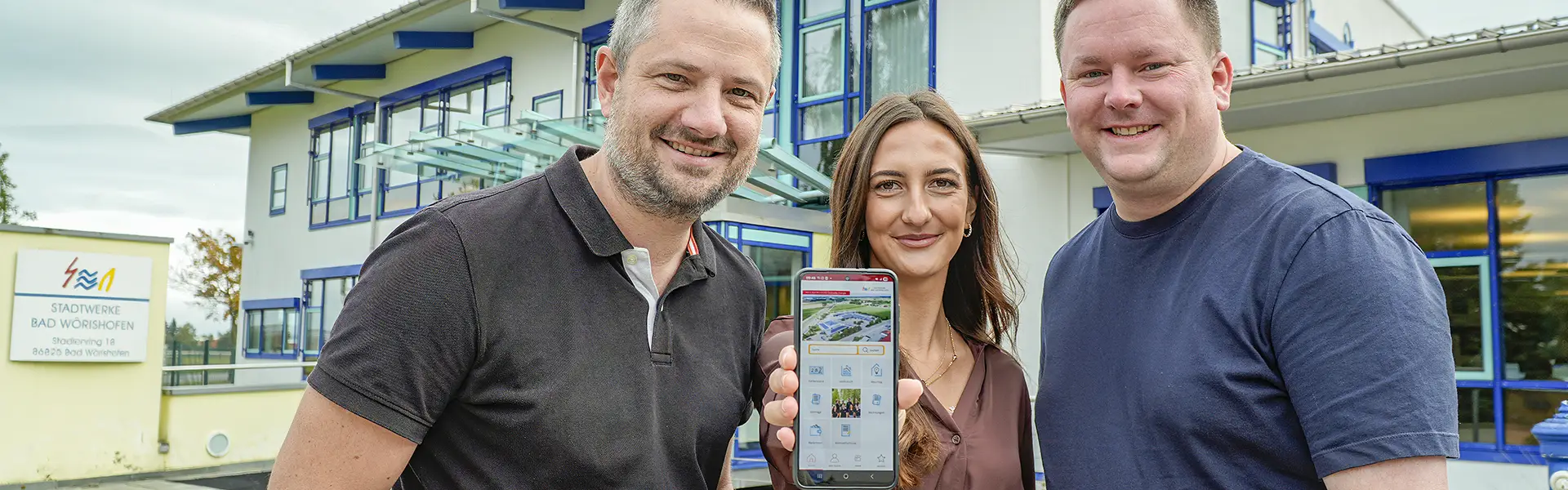 Drei Personen, die in die Kamera blicken. Die Person in der Mitte halt ein Smartphone in die Kamera.