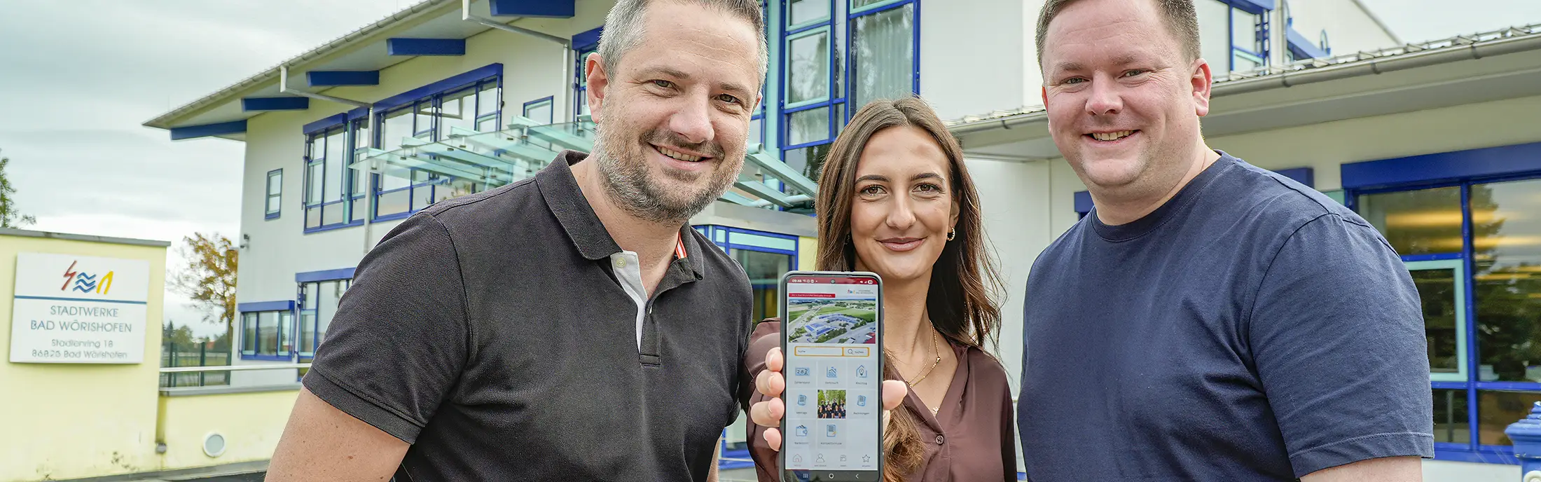 Drei Personen, die in die Kamera blicken. Die Person in der Mitte halt ein Smartphone in die Kamera.